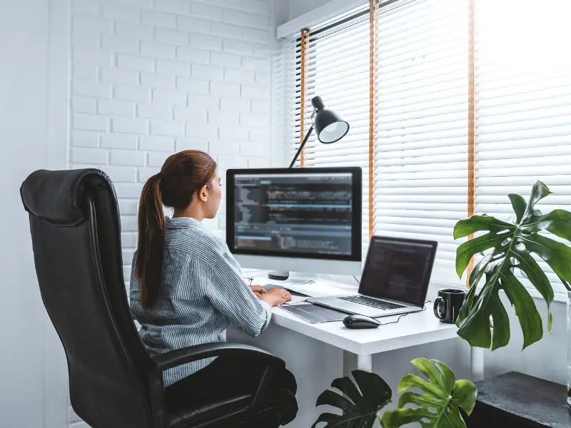 Arbeitsplatzgestaltung im Homeoffice. Junge Frau am ergonomischen Arbeitsplatz mit Laptop, Bildschirm, Maus, Lampe und Pflanzen