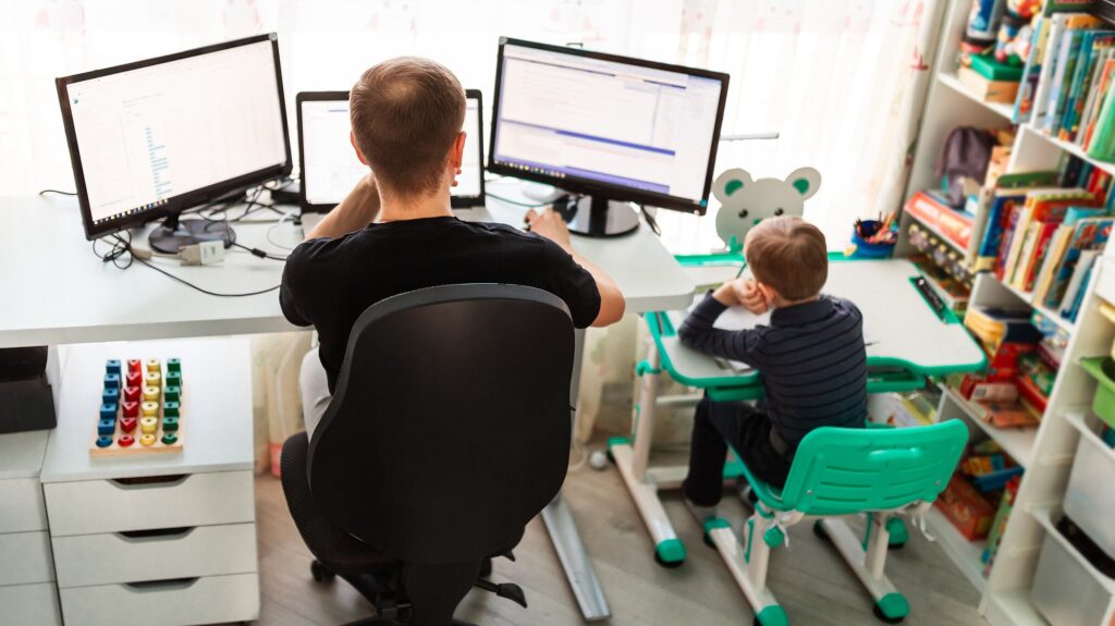 Ein Mann arbeitet von zu Hause aus an mehreren Monitoren, während ein Kind an einem kleinen Schreibtisch neben ihm sitzt. Der Raum ist hell und mit Büchern sowie Spielzeug ausgestattet, was eine familiäre und flexible Arbeitsumgebung zeigt.